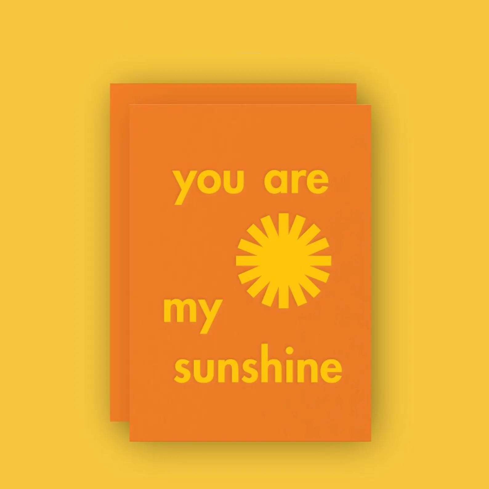 An orange greeting card with yellow foil-stamped text reads “you are my sunshine,” with a stylized sun icon replacing the word "sun." The card has a warm, cheerful design and comes with a matching orange envelope, set against a bright yellow background.