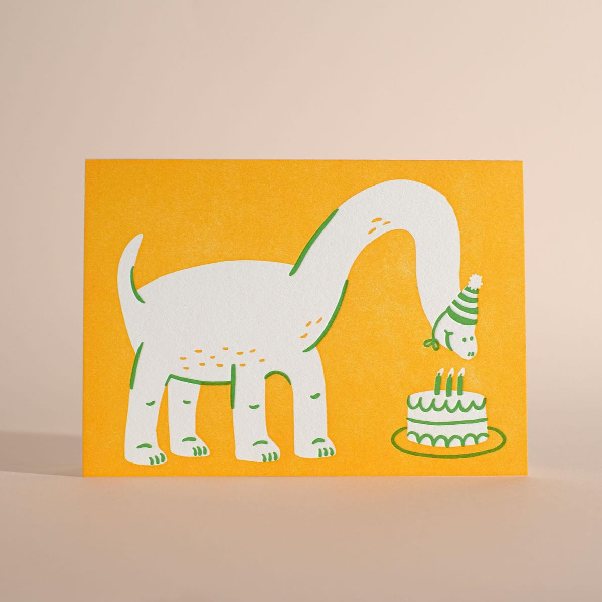 A bright yellow greeting card features a playful illustration of a dinosaur wearing a striped party hat. The dinosaur bends its long neck toward a birthday cake with three candles, creating a cheerful and whimsical birthday design.
