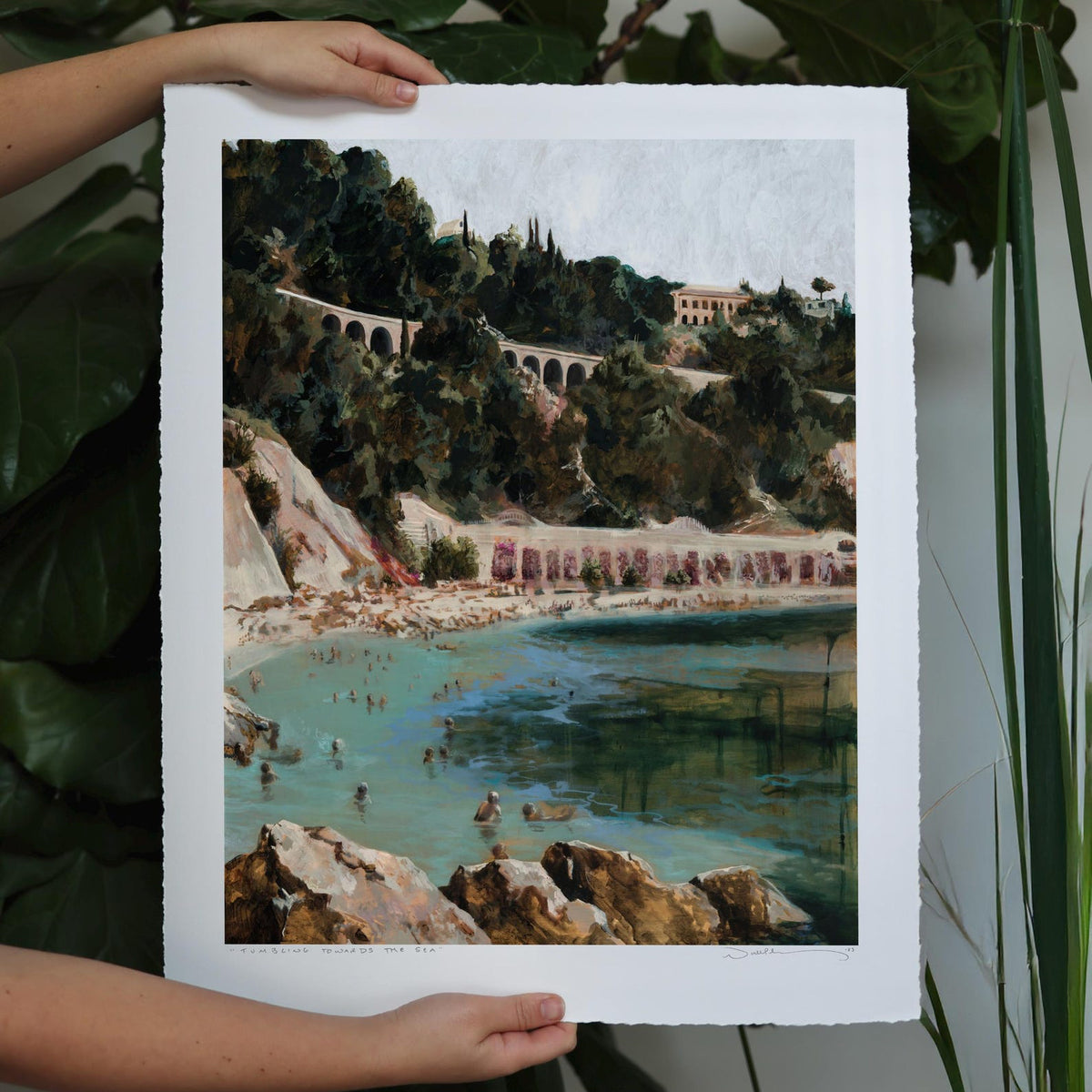 Lush greenery crawls around arched train tracks, tumbling towards serene blue waters with masses of people. 