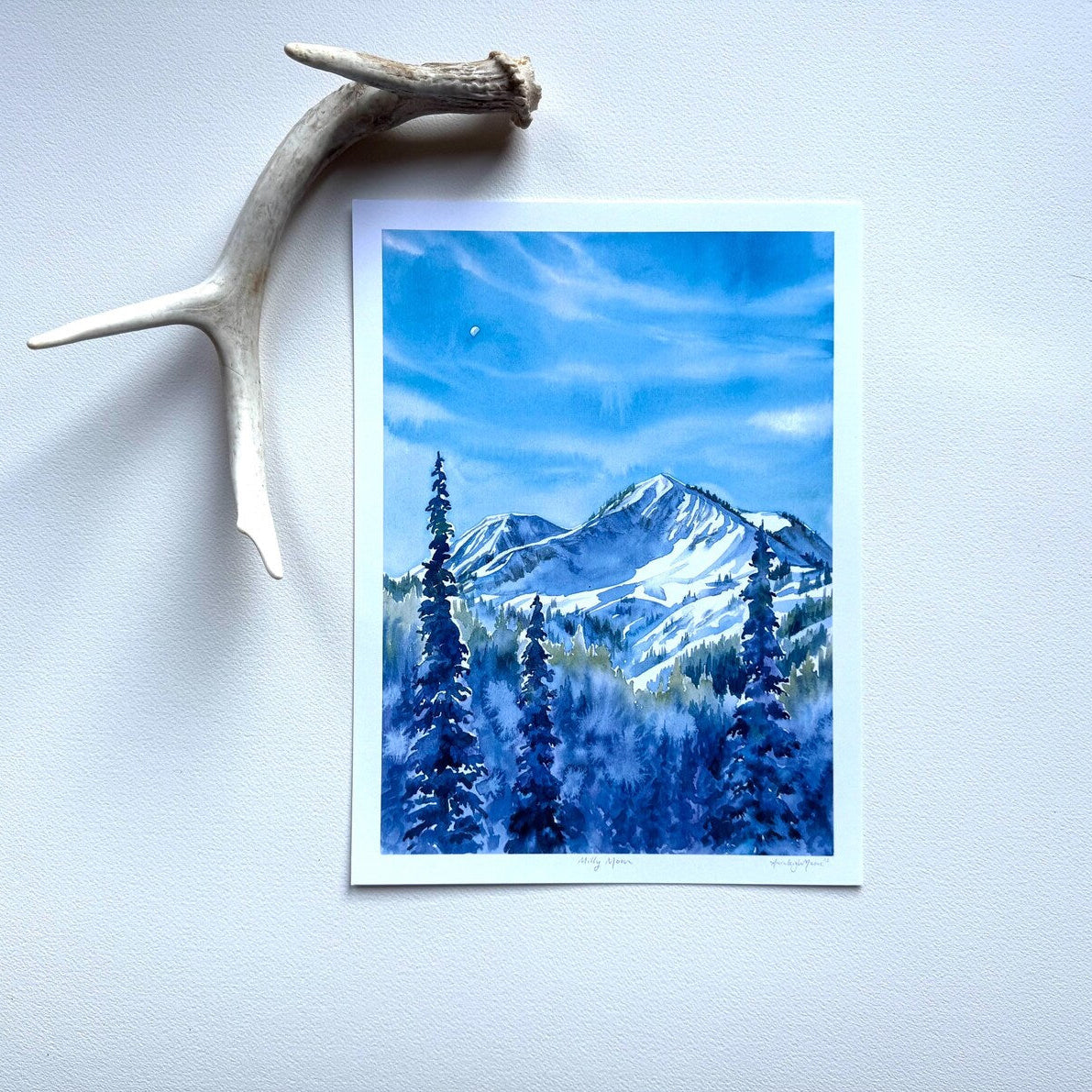 Vibrant blue watercolors create a magnificent mountain range with snowy pines in the forefront. Tiny moon is visible in the sky. Print is against a white wall with a decorative deer antler. 