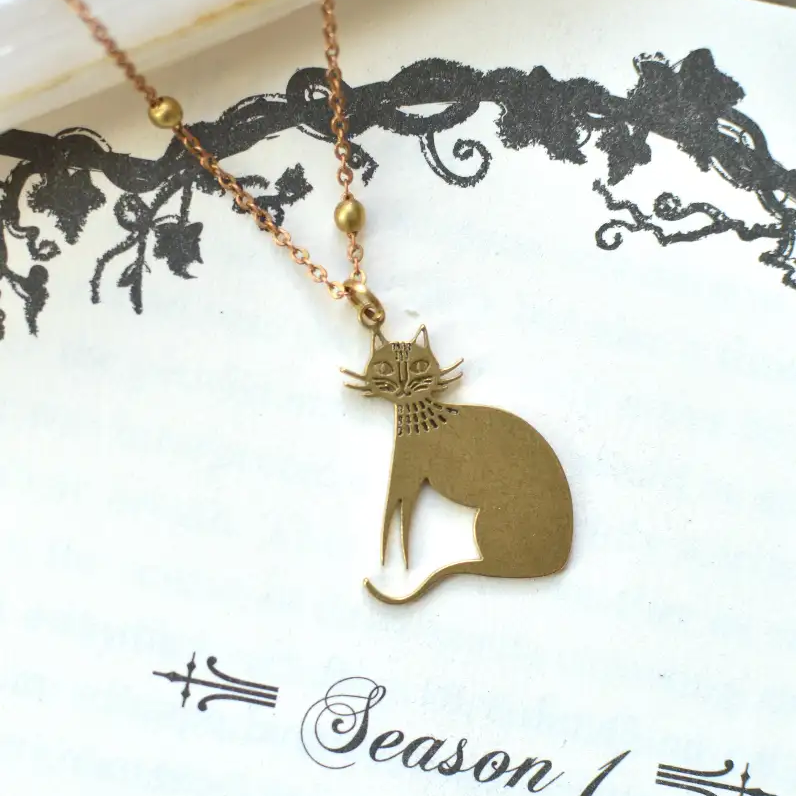 A gold-tone cat pendant necklace featuring a minimalist sitting cat silhouette with delicate facial details, paired with a fine gold chain accented by small round beads. The necklace is displayed on a decorative white card with black vintage-inspired floral artwork.