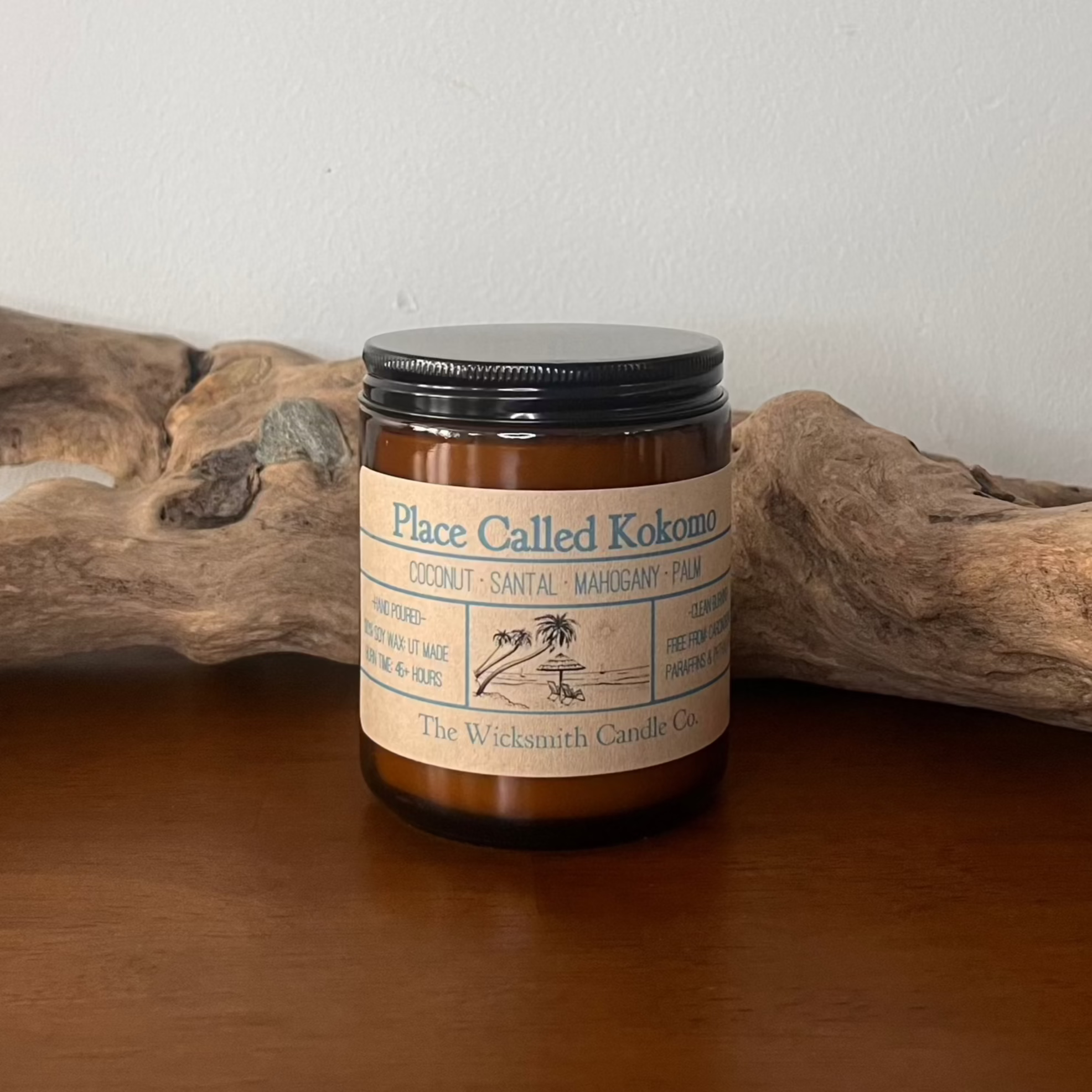 Amber glass candle jar labeled ‘Place Called Kokomo’ by The Wicksmith Candle Co., featuring scent notes of coconut, santal, mahogany, and palm, placed on a wooden surface with a piece of driftwood in the background