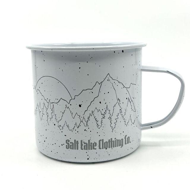 white enamel mug with black speckles, mountains, trees, and a sun
