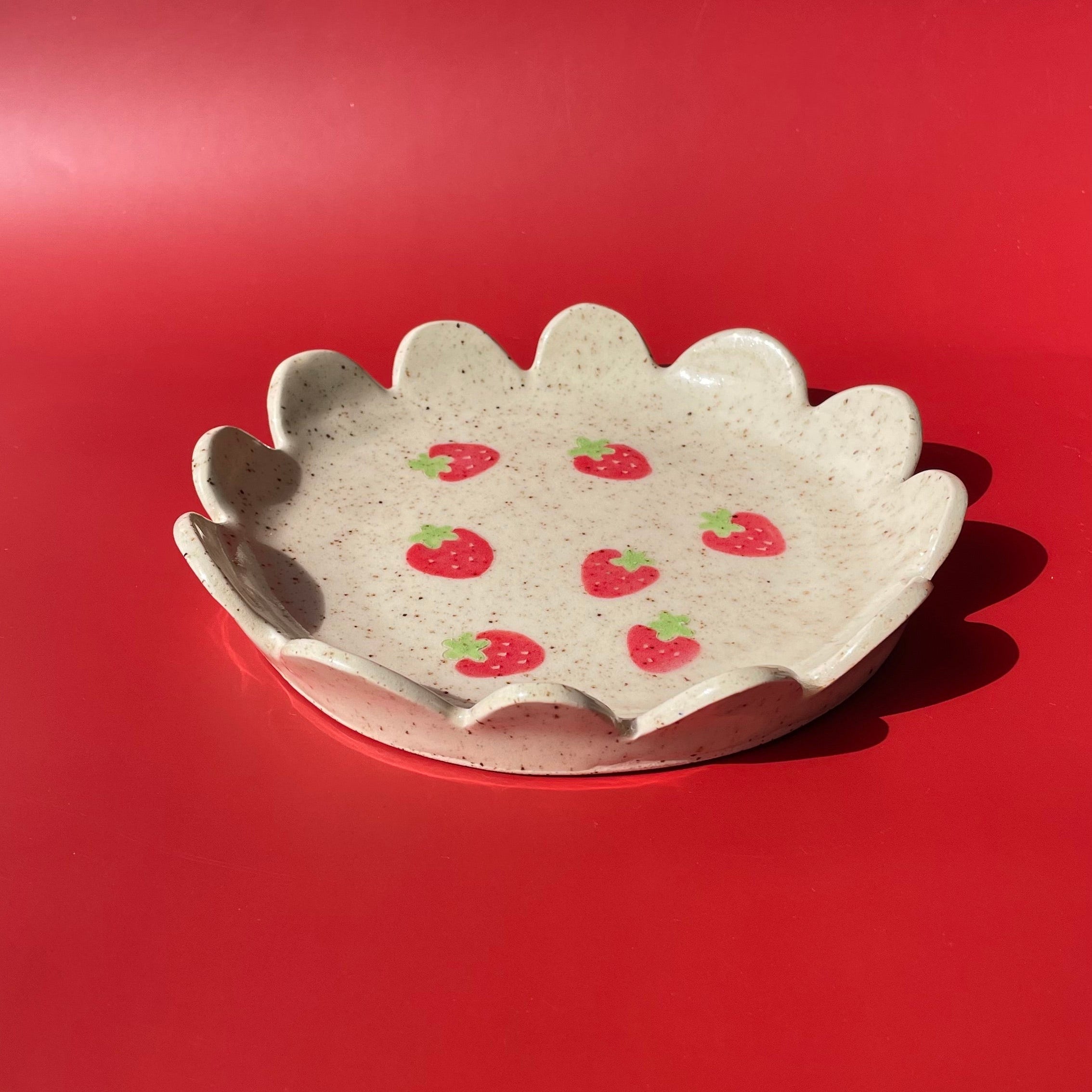 handmade ceramic dish with scalloped edges, finished with an off white glaze with brown specks, and red and green strawberry details painted in the center