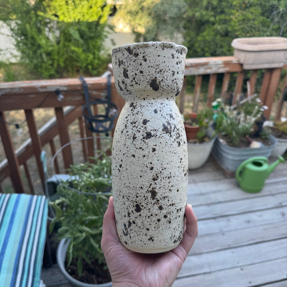 A hand holding a brown speckled vase in front of potted plants on a wooden deck outside. 