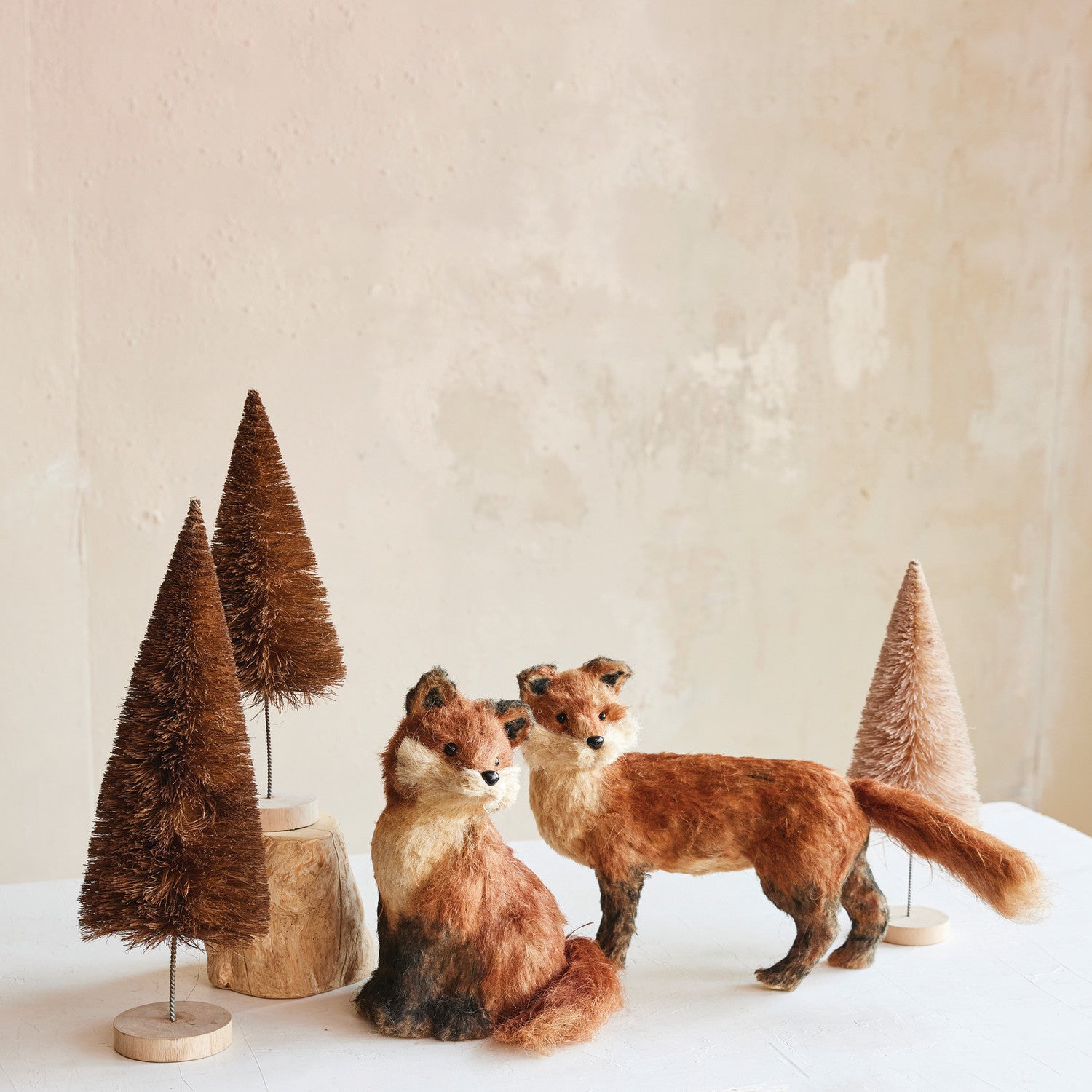 Two fox figurines standing next to two small brown trees on a white surface with a beige background.